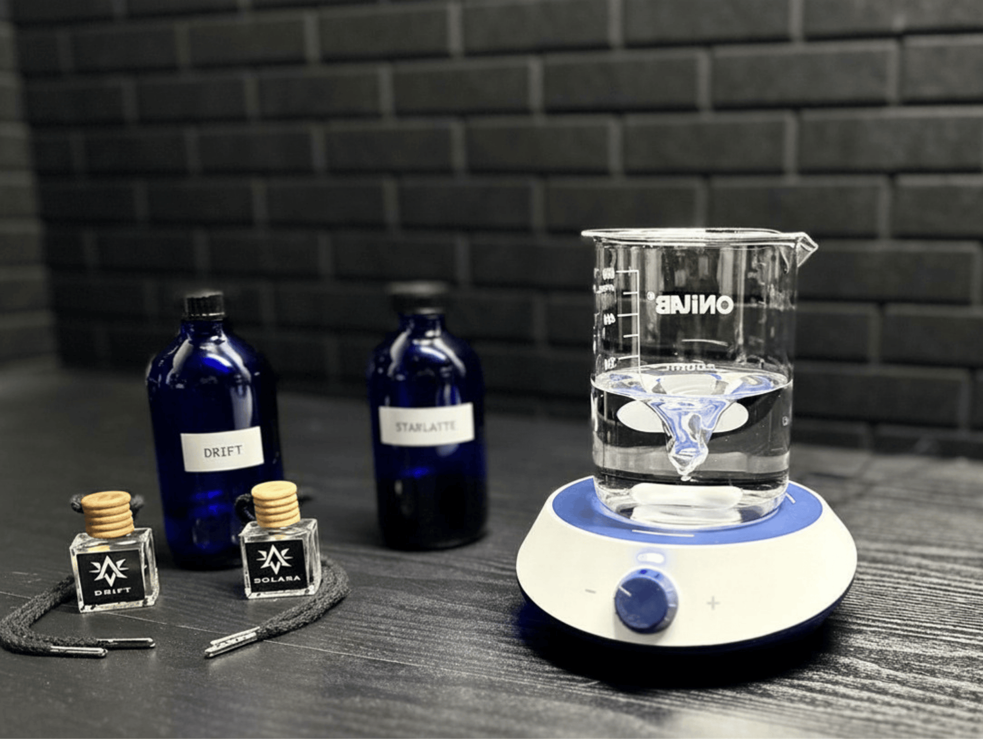 Starlite Avenue fragrance lab setup with magnetic stirrer, blue ingredient bottles, and Drift and Solara car air fresheners on a dark surface